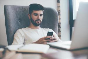 escribir un mensaje de negocios rápido. joven confiado sosteniendo un teléfono inteligente y mirándolo mientras estaba sentado en su lugar de trabajo en la oficina foto