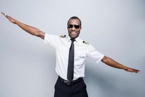 As a plane. Happy African pilot in uniform gesturing and smiling while standing against grey background photo