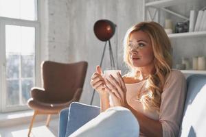 Just a minute to herself. Attractive young woman holding a cup and looking away with smile while sitting on the sofa at home photo