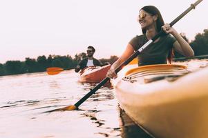 Enjoying nice time on river together. Low angle view of beautiful young couple kayaking on river together and smiling with sunset in the background photo