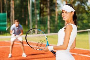 Feeling confident of winning. Beautiful young woman holding tennis racket and looking over shoulder with smile while man in sports clothing standing in the background photo