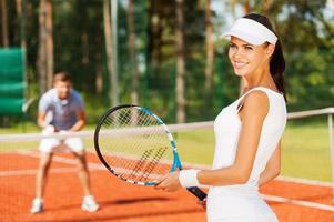 sentirse seguro de ganar. bella joven sosteniendo una raqueta de tenis y mirando por encima del hombro con una sonrisa mientras un hombre con ropa deportiva se para en el fondo foto