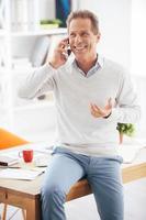 Good business talk. Cheerful mature man talking on the mobile phone and smiling while leaning at the desk in office photo