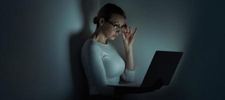 bella joven ajustándose las gafas mientras trabaja en una laptop contra una pared oscura foto