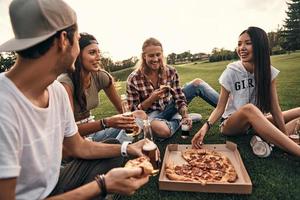 Fun time with friends. Group of young smiling people in casual wear enjoying pizza and beer while sitting on the grass outdoors photo