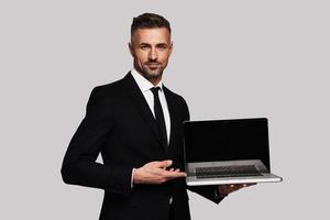 Look over here Handsome young man in full suit pointing copy space on laptop and smiling while standing against grey background photo