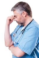 Depressed surgeon. Side view of depressed mature doctor touching his face with hand and keeping eyes closed while standing isolated on white photo