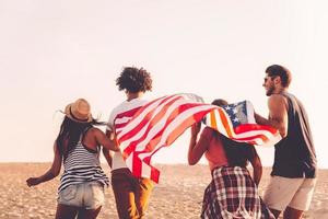 joven y libre. vista trasera de cuatro jóvenes que llevan la bandera americana mientras corren al aire libre foto