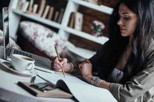 Putting ideas into something real. Thoughtful young woman writing something down in her notebook while sitting at her working place photo