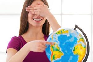 Choosing a traveling point. Cheerful little girl in formalwear examining globe with a loupe while sitting at the table photo