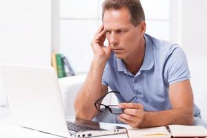 Thinking about solution. Thoughtful mature man looking at laptop and touching head with hand while sitting on the couch at home photo