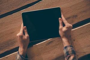 Living in digital age. Top view close-up image of man holding digital tablet while sitting at the rough wooden table photo