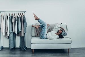 Lazy mood. Thoughtful young woman in casual wear touching her head with hand and putting feet up while lying on sofa at home near her clothes hanging on the racks photo