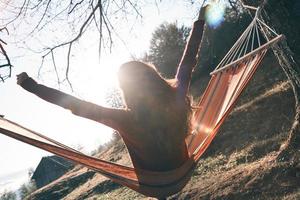 Enjoying the most beautiful place.  Rear view of young woman keeping arms outstretched while relaxing in hammock outdoors photo