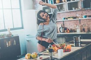 qué hermoso día hermosa joven mujer de raza mixta en auriculares cocinando ensalada y bailando mientras está de pie en la cocina en casa foto