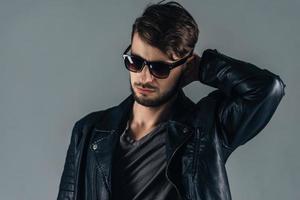 Trendy look. Confident young man in sunglasses looking down while standing against grey background photo