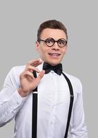 por favor, no un joven nerd aterrorizado con corbata de moño y tirantes haciendo una mueca y gesticulando mientras está de pie contra un fondo gris foto