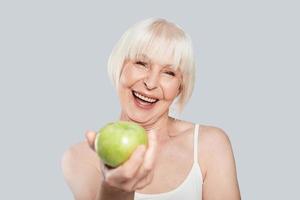 Would you like some Beautiful senior woman holding an apple and smiling while standing against grey background photo