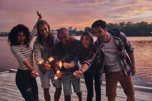 Friends make each other happy. Group of young people in casual wear smiling and holding sparkers while standing on the pier photo