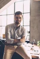 Cheerful businessman. Confident young man in smart casual wear keeping arms crossed and looking at camera with smile while leaning at the desk in office photo