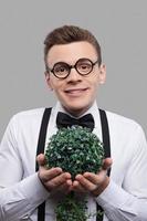 Young botanist. Portrait of cheerful young man in bow tie and suspenders holding a plant in his hands and smiling while standing against grey background photo