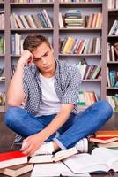 Tired of studying. Frustrated young man touching his head with hand and looking at books while sitting against bookshelf photo