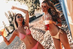 sintiéndose geniales dos mujeres jóvenes atractivas sonriendo y disfrutando de cócteles mientras se divierten en la playa foto