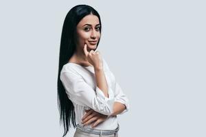 Thinking about new business aproaches. Confident young woman in white shirt smiling and looking at camera while standing against grey background photo