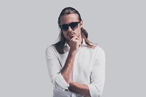 Thoughtful handsome. Handsome young man in white shirt and sunglasses looking at camera and holding hand on chin while standing against grey background photo