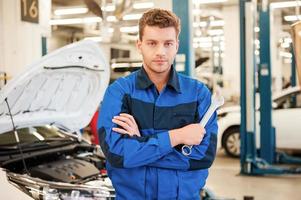 Confident expert. Confident young man holding a wrench and looking at camera while standing in workshop with car in the background photo