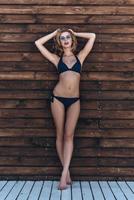 joven y hermoso. toda la longitud de una joven atractiva en bikini manteniendo las manos detrás de la cabeza mientras posa contra la pared de madera al aire libre foto