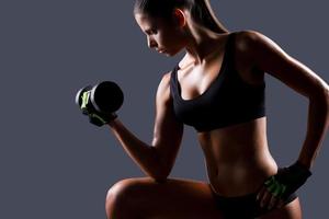 Strengthening her muscles. Side view of beautiful young woman exercising with dumbbells while standing against grey background photo