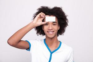 Copy space on my forehead. Cheerful African teenager holding plastic card at his forehead and looking at camera while standing isolated on grey background photo