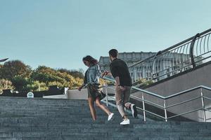 joven y lleno de energía. vista trasera completa de un hombre guapo y una mujer joven y atractiva tomándose de la mano mientras subía las escaleras al aire libre foto