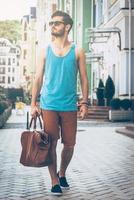 Exploring city. Full length of handsome young man in casual wear carrying bag and looking away while walking along the street photo