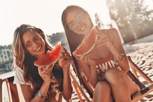 Simply having fun. Two attractive young women smiling and eating watermelon while sitting on the beach photo