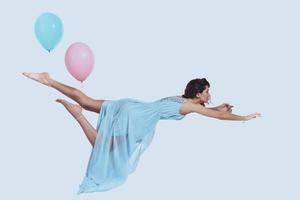 Beauty in mid-air. Studio shot of attractive young woman in elegant dress keeping arms outstretched while hovering against grey background photo