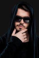 He got dark side. Confident young man in black hood holding hand on chin and looking at camera while standing against black background photo