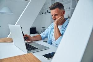 Tired mature man working on laptop while sitting at the office desk photo