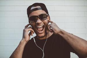 Loving this track Happy young African man adjusting his headphones and keeping mouth open while standing against the stoned wall outdoors photo