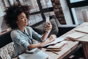 Answering important messages. Beautiful young African woman looking at smart phone and smiling while sitting at her working place in restaurant photo