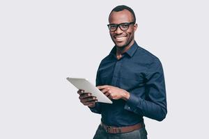 How may I help you Handsome young African man holding digital tablet and looking at camera with smile while standing against grey background photo