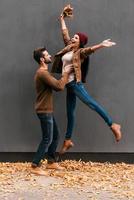 Autumn fun. Beautiful young couple having fun together while standing against grey wall with orange fallen leaves laying around them photo