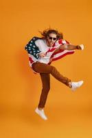Confident young man in casual clothing making a face and punching while hovering covered in American flag against yellow background photo