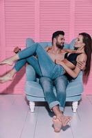 Happy to be in love. Handsome shirtless man carrying his girlfriend and smiling while sitting in the armchair against pink background photo