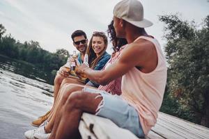 Enjoying every minute together. Group of happy young people in casual wear smiling and drinking beer while sitting on the pier photo