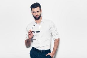 Handsome and stylish. Handsome young man in white shirt holding sunglasses and looking at camera while standing against white background photo