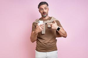 Surprised mature man pointing his smart phone while standing against pink background photo