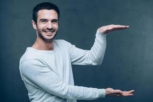 Copy space in his hands. Handsome young man holding copy space and looking at camera with smile while standing against grey background photo