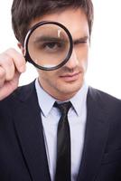 Businessman examining you. Serious young man in formalwear examining you with magnifying glass while standing isolated on white background photo